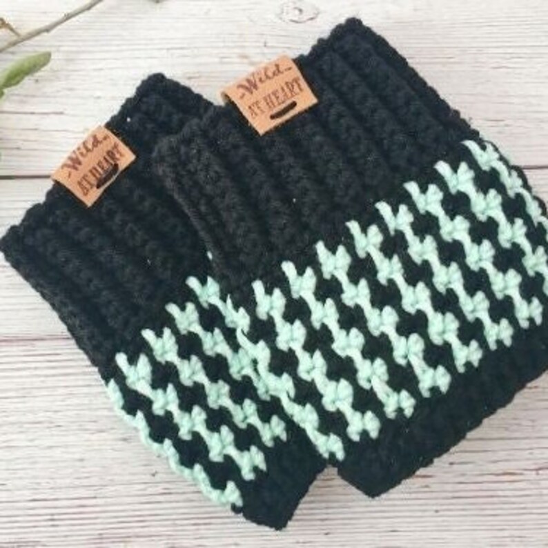 May include: A pair of black and mint green crochet leg warmers. The leg warmers feature a houndstooth pattern in mint green on the lower half and solid black on the upper half. Each leg warmer has a small, rectangular tag that reads "Wild at Heart".