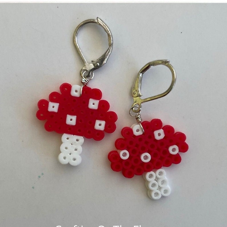 May include: A pair of handmade mushroom earrings. Each earring features a red mushroom cap with white spots and a white stem. The earrings are made from small, colorful beads and have silver-colored leverback closures.