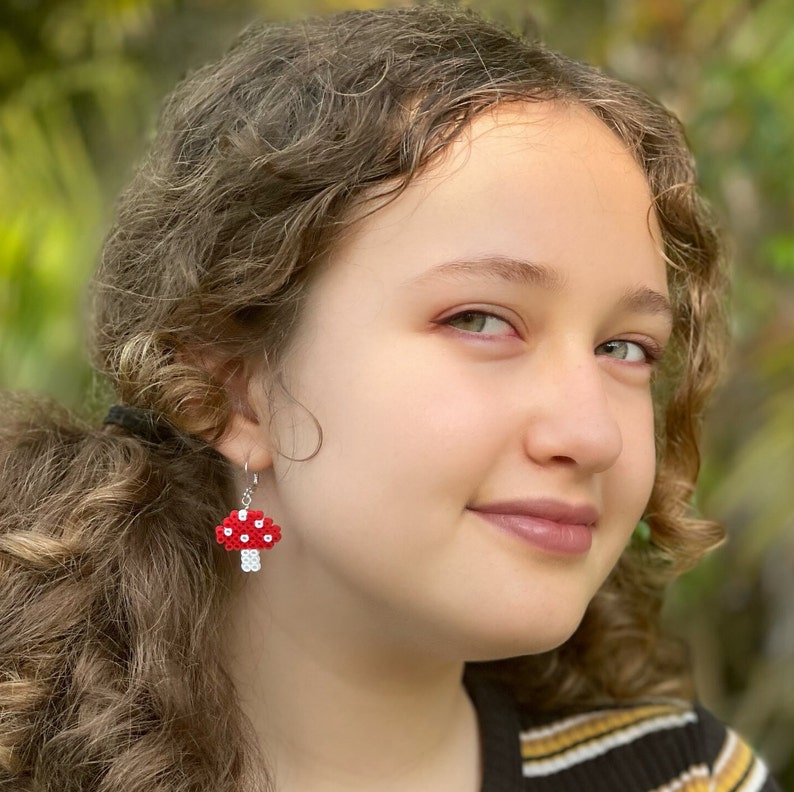 May include: Red and white mushroom earrings. The earrings are made of small beads and have a silver hook. The mushroom caps are red with white dots, and the stems are white. The earrings are worn by a person with curly brown hair.