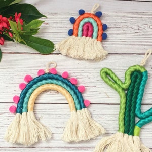 Op de afbeelding: Drie kleurrijke macramé wandhangers, elk met een ander ontwerp. De eerste is een regenboog met roze, blauwe en groene draad, de tweede is een regenboog met oranje, roze en blauwe draad, en de derde is een groene cactus.