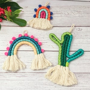 May include: Three colorful macrame wall hangings, each with a different design. The first is a rainbow with pink pom-poms, the second is a rainbow with blue pom-poms, and the third is a green cactus.