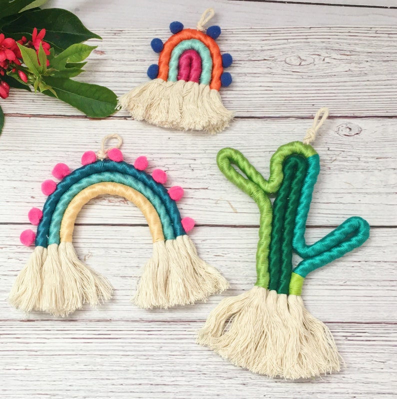 May include: Three colorful macrame wall hangings, each with a different design. The first is a rainbow with pink pom-poms, the second is a rainbow with blue pom-poms, and the third is a green cactus.
