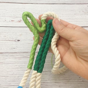 May include: A close-up of a white rope with green and dark green yarn wrapped around it. The yarn is twisted and knotted, creating a decorative effect.