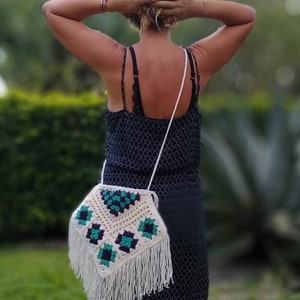May include: A white crocheted bag with green and black geometric patterns and fringe. The bag is worn over the shoulder of a person wearing a black dress.