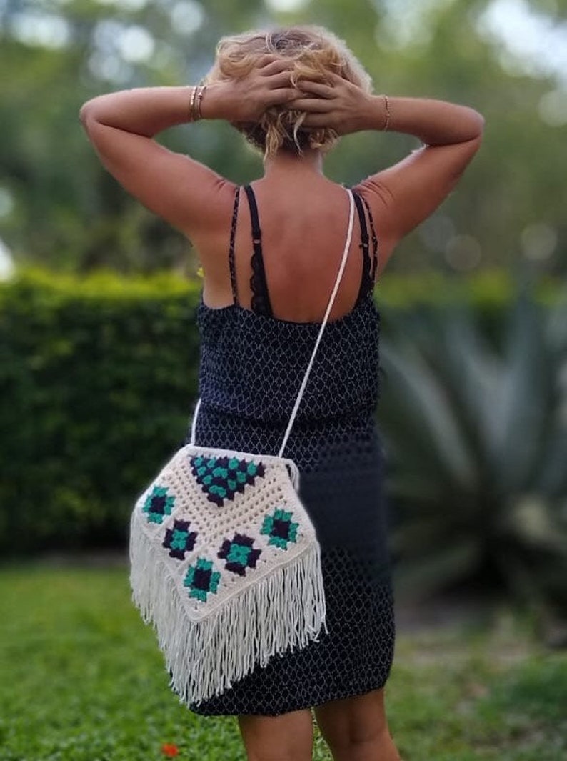 May include: A white crocheted bag with green and black geometric patterns and fringe. The bag is worn over the shoulder of a person wearing a black dress.