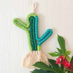Op de afbeelding: Een groene en blauwe macramé cactus wandhanger met een witte kwast.