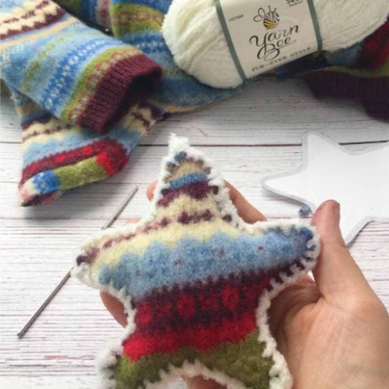 Op de afbeelding: Een stervormige ornament gemaakt van een kleurrijke gebreide stof met een witte rand. De ornament wordt in een hand gehouden.