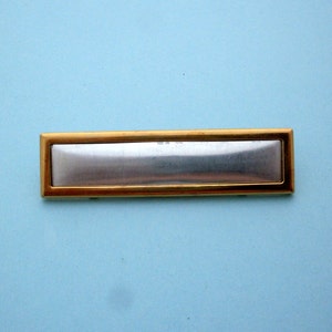 May include: A gold-colored metal name tag with a silver-colored rectangular insert. The tag is blank.
