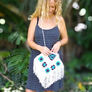 May include: A woman wearing a black and white patterned dress holds a white crocheted bag with turquoise and purple accents. The bag has fringe detailing.