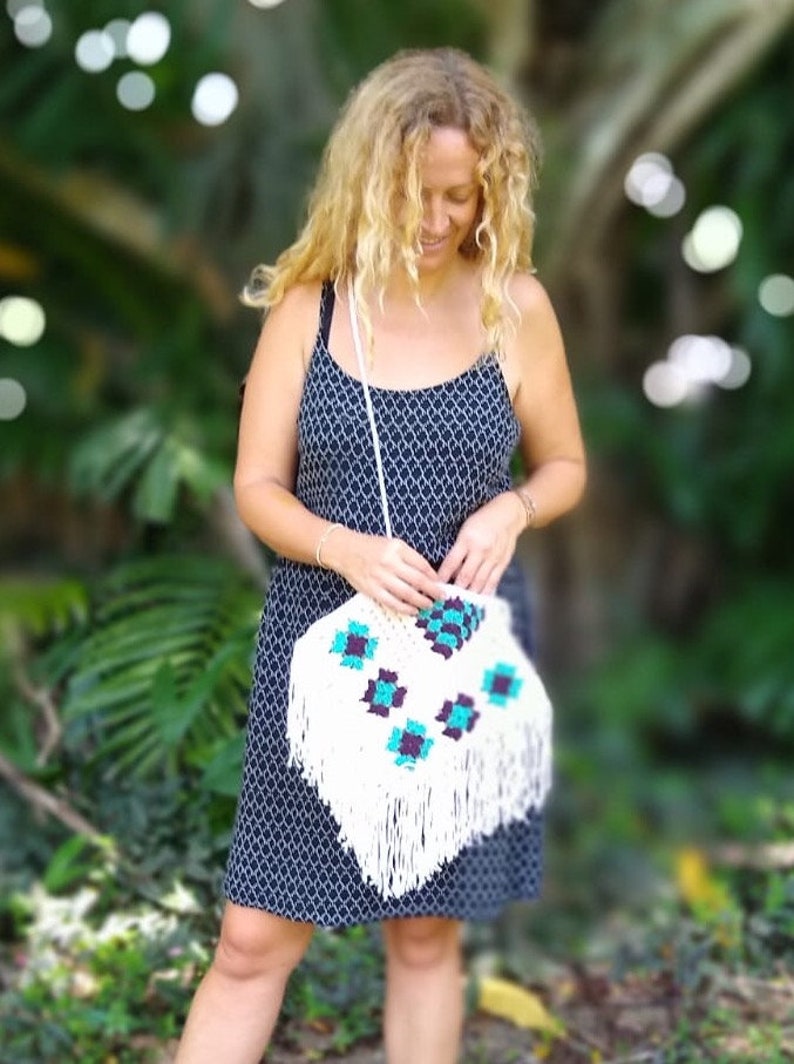 May include: A woman wearing a black and white patterned dress holds a white crocheted bag with turquoise and purple accents. The bag has fringe detailing.