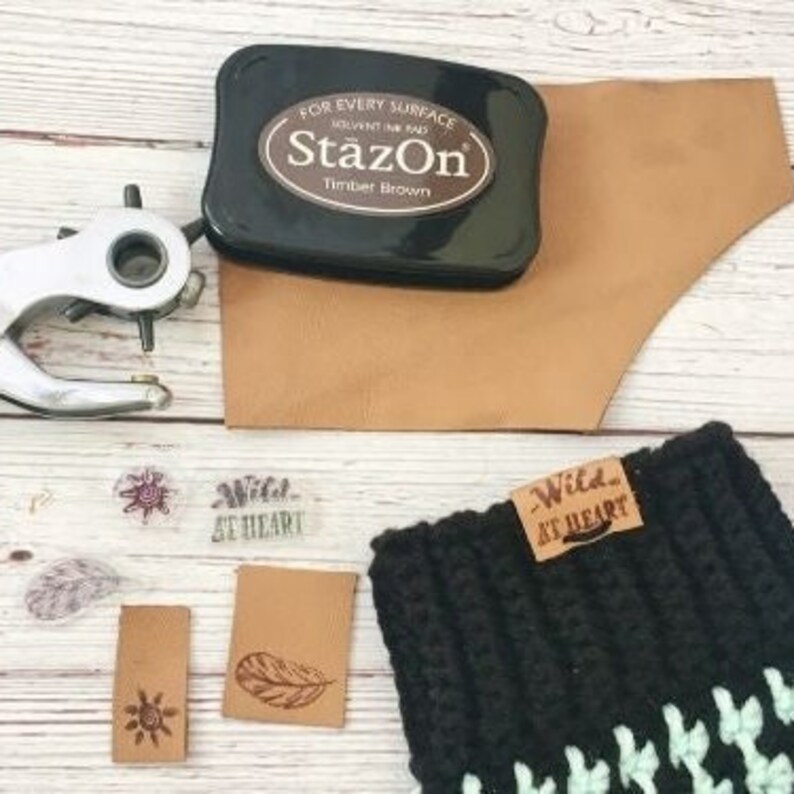 May include: An assortment of leather crafting supplies, including a black StazOn Timber Brown ink pad, a leather punch, and various leather tags. One tag is stamped with "Wild at Heart" and a feather design. A black and green crocheted item is also present.