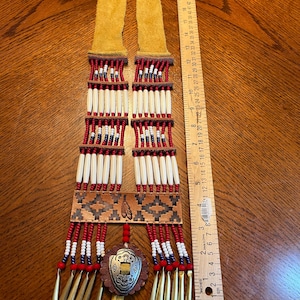May include: A Native American-style necklace with a tan suede neck strap. The necklace features rows of white, red, and blue beads, with a central pendant of a shield design. The necklace is approximately 14 inches long.