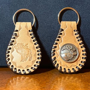 May include: Two leather keychains with a brown braided border. One keychain features a moose design and the other features an elk design. Both keychains have a silver metal button with the animal design.