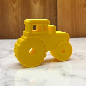 Könnte beinhalten: Ein hellgelber Spielzeugtraktor mit großen, kreisförmigen Rädern und einer rechteckigen Kabine. Das Spielzeug besteht aus einem festen Material und steht auf einer weißen Marmoroberfläche mit einem hölzernen Hintergrund.