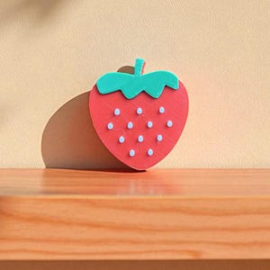 May include: A red strawberry-shaped object with a green stem and small white dots. The object is placed on a wooden surface, casting a shadow against a beige background. The strawberry design is simple and stylized.