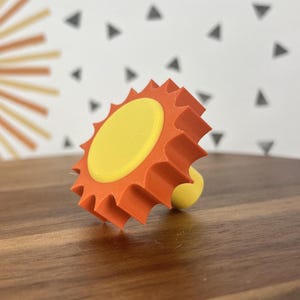 May include: A decorative bottle stopper in the shape of a sun. It features a yellow central disc and an orange, spiky surround. The stem is a small, rounded yellow shape. The background has a sun design and a pattern of grey triangles.