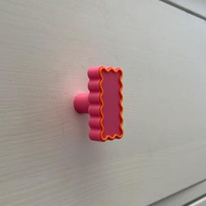 May include: A pink cabinet knob with a rectangular shape and wavy edges, outlined in orange. The knob is mounted on a white wooden surface, providing a pop of color and a playful design element for furniture.