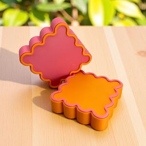 Peut inclure: Deux dessous de verre colorés aux bords ondulés. L'un est orange vif avec un bord rose, et l'autre est rose avec un bord orange. Les dessous de verre sont sur une surface en bois clair, avec un arrière-plan vert flou.