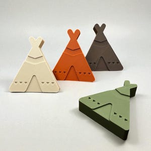 May include: Four small, colourful teepee-shaped decorative objects. One is cream, one is orange, one is brown, and one is green. Each has a cutout design and a small, heart-shaped top. The objects are arranged on a white surface.