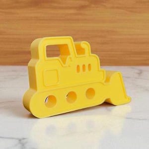 May include: A bright yellow toy bulldozer with a blade and track wheels. The toy is made of a solid material and has a rectangular cab with a window. The bulldozer is a construction-themed toy.