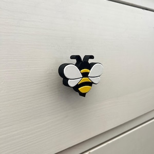May include: A black, yellow, and white bee-shaped cabinet knob. The knob has a 3D design with a black body, white wings, and yellow and black stripes on the abdomen. The knob is mounted on a white wooden surface.