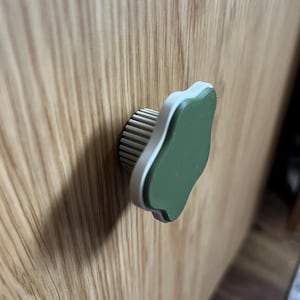 May include: A cabinet knob with a leaf-like design. The knob features a light green top with a white border and a ribbed, beige base. It is mounted on a wooden cabinet door.