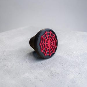 May include: A circular black knob featuring a raised red spider web design on its flat front face. The knob has a tapered cylindrical body with a smooth matte finish, providing a distinct contrast between the black base and the vibrant red web pattern.