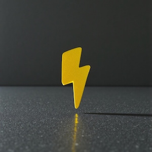 May include: A bright yellow lightning bolt shape stands on a dark gray surface, casting a shadow. The background is a darker gray. The bolt is angled slightly to the right, with a textured surface.