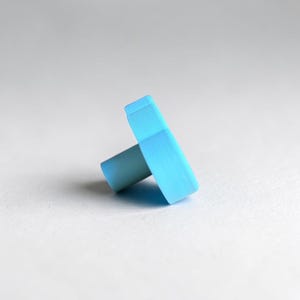 May include: A light blue cabinet knob with a flat, rectangular front face and a cylindrical base. The surface of the knob has a smooth, matte finish with visible horizontal texture lines across the front, providing a consistent solid color.
