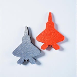 Könnte beinhalten: Zwei Flugzeug-förmige Objekte. Eines ist silberfarben, das andere leuchtend orange. Beide haben eine strukturierte Oberfläche und sind vor einem weißen Hintergrund platziert. Die Objekte sind wahrscheinlich dekorativ oder Spielzeug.