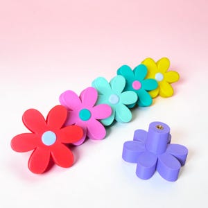 Könnte beinhalten: Eine Sammlung von sechs blumenförmigen Schubladenknöpfen in leuchtenden Farben wie Rot, Pink, Hellblau, Türkis, Gelb und Lila. Jeder Knopf verfügt über eine kontrastierende kreisförmige Mitte und einen metallenen Gewindeeinsatz auf der Rückseite zur Montage.