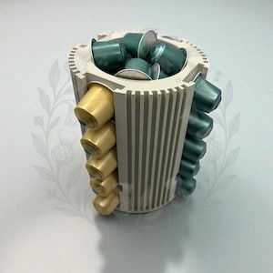 Könnte beinhalten: Ein zylindrischer, cremefarbener Kaffeepadhalter, gefüllt mit grünen und goldenen Kaffeepads. Der Halter hat eine geriffelte Außenseite und eine kreisförmige Oberseite. Die Pads sind in Reihen angeordnet.