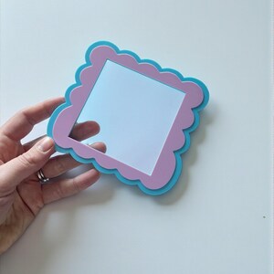 May include: A square-shaped hand mirror with a scalloped edge. The mirror has a light blue frame with a pink scalloped border and a reflective surface. The mirror is held by a hand.