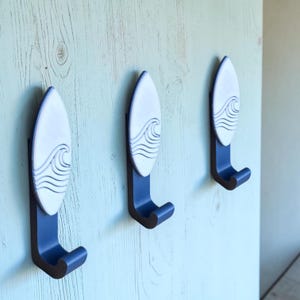 May include: Three wall-mounted hooks, each featuring a vertical surfboard-shaped plaque in white with a raised, dark blue wave design. The hooks themselves are dark blue, extending outward from the base of each surfboard-shaped plaque.