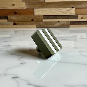 May include: An olive green cabinet knob with two white stripes. The knob has a rounded base. The item is set against a marble surface with a wooden plank background.
