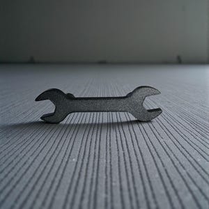 Könnte beinhalten: Ein kleiner, schwarzer Schraubenschlüssel mit doppeltem Ende. Der Schraubenschlüssel besteht aus Metall und liegt auf einer strukturierten, grauen Oberfläche. Das Werkzeug dient zum Anziehen oder Lösen von Muttern und Bolzen.