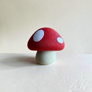 May include: A small mushroom figurine featuring a textured red cap with two smooth white circular spots and a pale green stem. The surface of the object displays visible concentric circular lines from the manufacturing process.