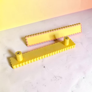 May include: Two rectangular drawer pulls with a scalloped edge design. The pulls are yellow with a thin pink accent line along the scalloped border. Each pull features two cylindrical mounting posts with metal inserts for screw attachment.