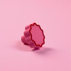 May include: A pink, flower-shaped cabinet knob with a rounded stem. The knob has a scalloped edge with a darker pink trim. The background is a solid pink color, creating a monochromatic aesthetic. This decorative knob is suitable for furniture.