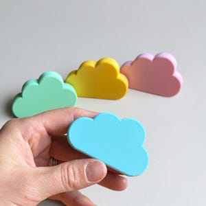 May include: A set of four cloud-shaped objects in pastel colors including mint green, yellow, light pink, and bright blue. Each cloud has a smooth, matte finish and a rounded, scalloped top edge. One blue cloud is held in the foreground.
