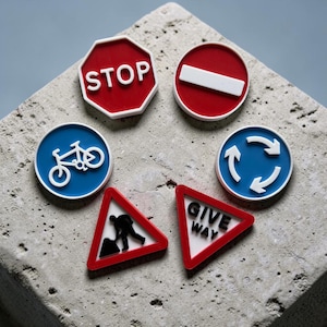 May include: A collection of six small, flat, circular, octagonal, and triangular road sign replicas. The set includes a red stop sign, a red no entry sign, a blue bicycle sign, a blue roundabout sign, a red road work sign, and a red give way sign.