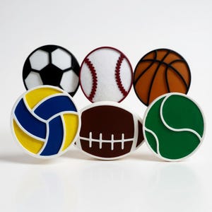 May include: A collection of sports-themed decorative knobs. The knobs feature designs of a soccer ball, baseball, basketball, volleyball, football, and tennis ball. Each knob has a white border and is set against a white background.