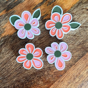 May include: Four flower-shaped embellishments with a retro design. Each flower has pink and orange petals with a white outline and a green centre. Green leaves are attached to some of the flowers. The flowers are on a wooden surface.