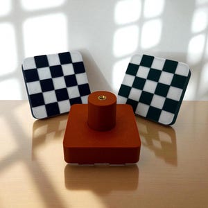 Könnte beinhalten: Ein Set aus drei Artikeln: eine quadratische, rotbraune Basis mit zylindrischer Oberseite und zwei quadratische Untersetzer mit Schachbrettmustern in Schwarz und Weiß sowie Grün und Weiß. Die Artikel sind auf einer hellen Oberfläche angeordnet.