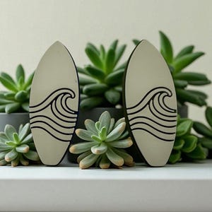 Peut inclure: Deux décorations en forme de planche de surf avec un motif de vagues noir. Les planches de surf sont beige clair avec un bord noir. Le fond présente des succulentes vertes dans des pots gris.