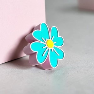 May include: A flower-shaped charm featuring four bright turquoise petals with a solid yellow center. The charm has a thick, light lavender base and a white outline surrounding each petal, creating a distinct layered appearance and a smooth finish.