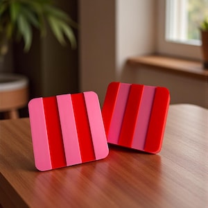 May include: Two square coasters with a striped design. Each coaster features alternating vertical stripes of red and pink. The coasters are made of a glossy material and sit on a wooden surface.