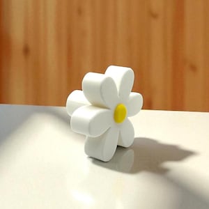 Peut inclure: Un objet blanc en forme de marguerite avec un centre jaune. La fleur a six pétales arrondis et est posée sur une surface claire avec un fond en bois.