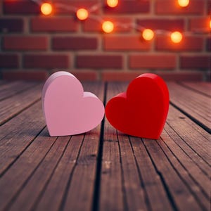 May include: Two heart-shaped objects, one pink and one red, sit on a wooden surface. The background features a brick wall and a string of glowing lights, creating a romantic ambiance. The hearts are the focal point, symbolizing love and affection.