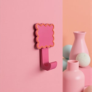 Peut inclure: Un crochet mural rose avec un dessus festonné et un contour jaune. Le crochet est monté sur un mur rose. Des vases et des sphères roses décoratifs sont à l'arrière-plan.